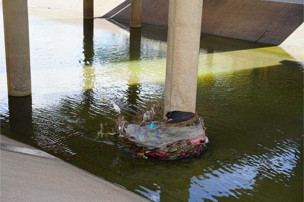Sous un pont, les pieds des colonnes sont immergés dans l'eau. Des débris, des déchets et des éléments polluants sont accrochés à l'une des colonnes, illustrant les effets de la montée des eaux sur l'environnement et la pollution causée par une crue.